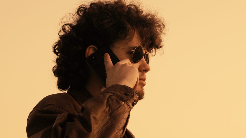 Curly-haired man in sunglasses on a phone call against a pale gold sky
