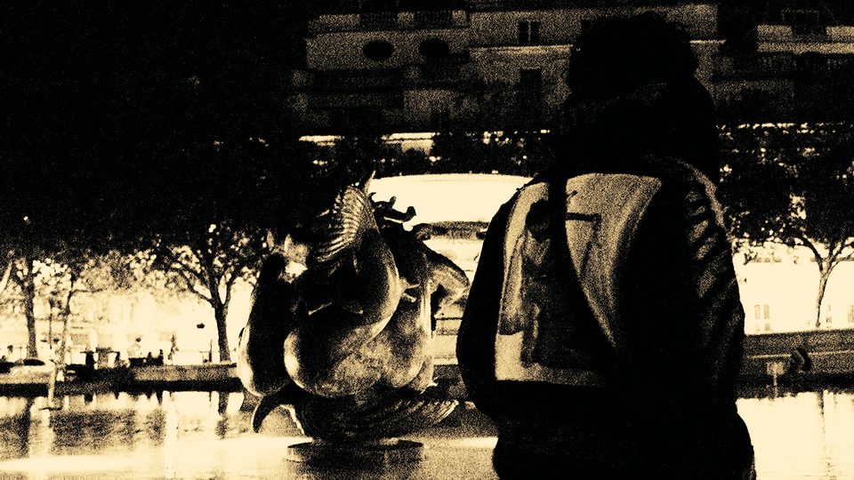 Black-and-white frame of a figure beside a fountain sculpture